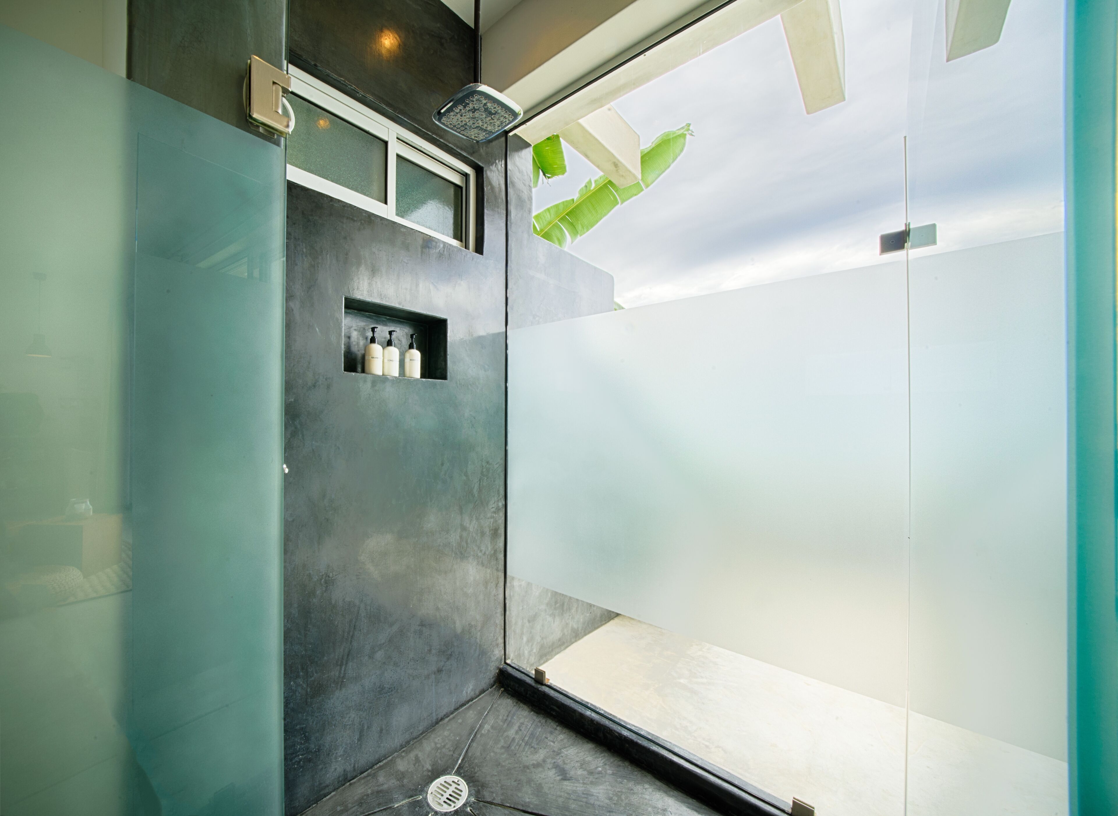 A modern bathroom with a shower, a small shelf with toiletries, and a view of the sky through a frosted glass door.