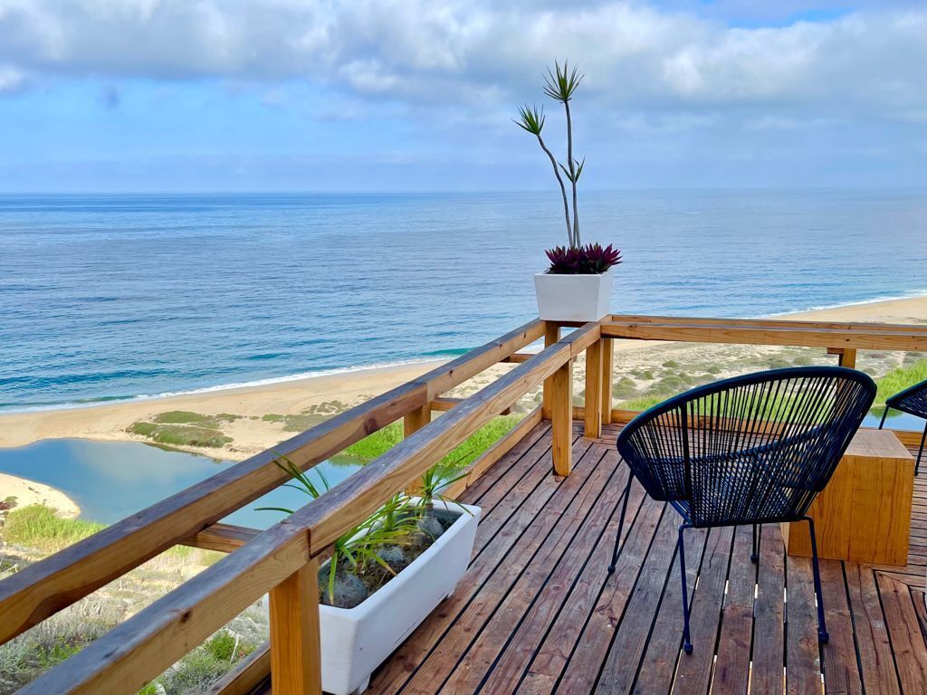 Um deck de madeira com guarda-corpo, vista para a praia e oceano. Há duas cadeiras e um vaso com plantas.