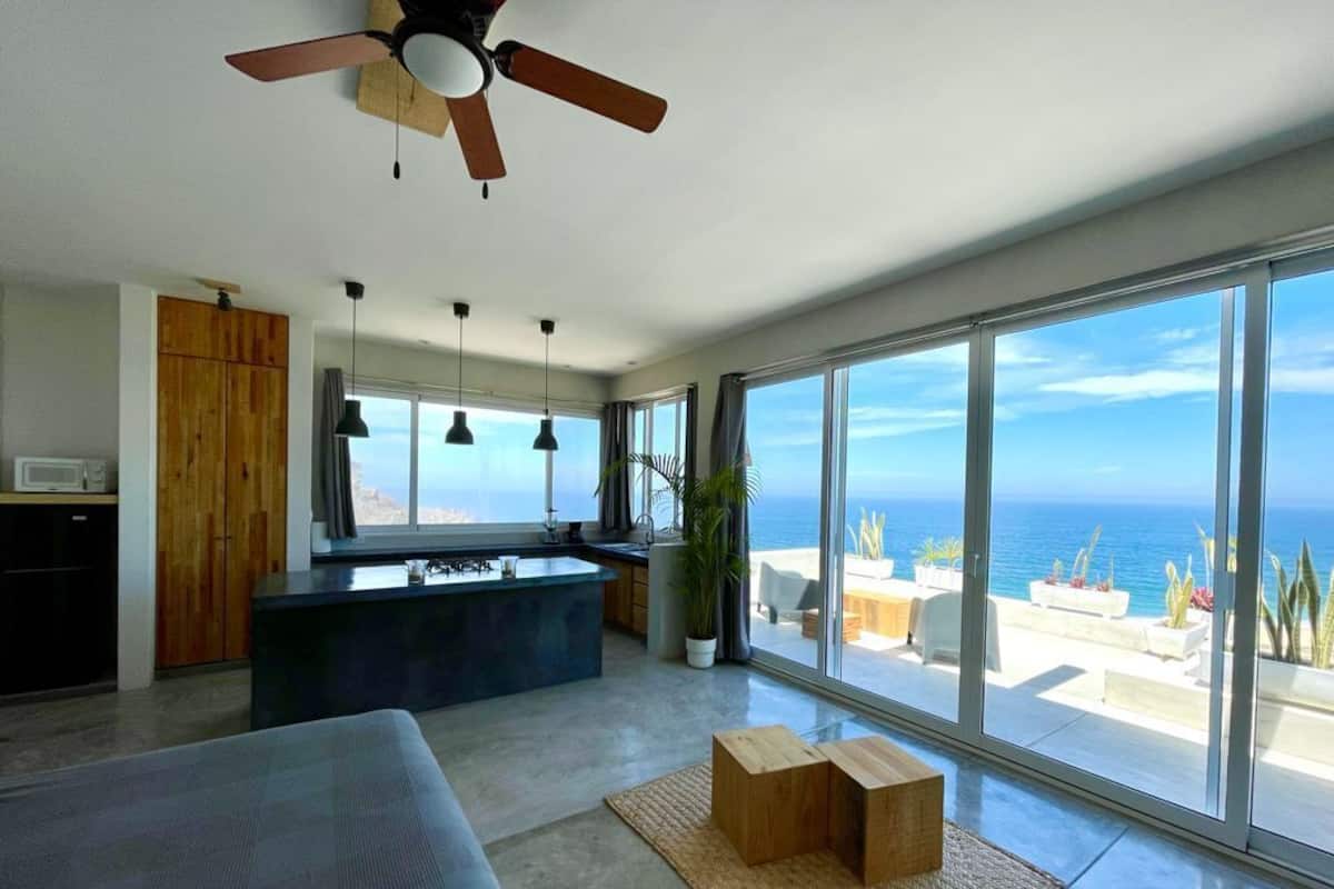 A modern kitchen with a large island, a ceiling fan, and a view of the ocean through large windows.
