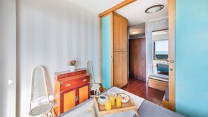 A bedroom with a bed, a wooden dresser, an ironing board, a small table with breakfast items, and a view of the outdoors. - La Brise Apartment by Wonderful Italy (Sanremo)