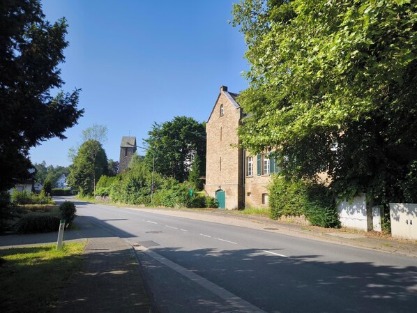 A quiet street with a prominent stone building, a clear sky, and lush greenery. - Cozy apartment in the Bergisches Land. Pet friendly! (Much)
