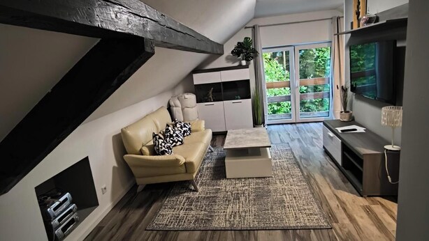 A modern loft-style living room with a sloped ceiling, built-in entertainment unit, and a large window offering a view of greenery.