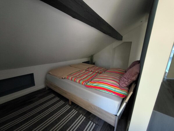 Une petite chambre avec un lit simple, un plafond en pente, un tapis à rayures et un mur uni.