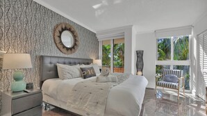 A modern bedroom with a large bed, a nightstand, a chair, and a view of the outdoors through a window. - Treetop Retreat Cozy 2 Bedroom in Grove (Miami)