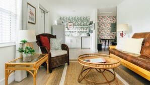 A living room with a brown leather sofa, a wicker chair, a small table with a lamp, and a kitchen in the background.