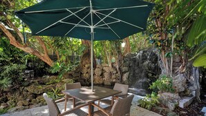 Une terrasse avec une table et des chaises sous un parasol vert, entourée d’une végétation luxuriante et d’une petite cascade.