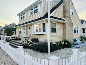 A two-story house with a white picket fence, a front porch with chairs, and a gravel driveway.