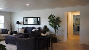 A modern living room with a dark sofa set, a coffee table, and a large mirror on the wall. There is a potted plant and a small table with decorative items. - Spacious Ranch w/Hot Tub & Game Room, Sleeps 14! (Greeley)
