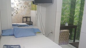 A bedroom with a bed, a TV mounted on the wall, a small table with a chair, and a view of greenery through a window. - CASADALUCIA  POUSADA (Salvador)