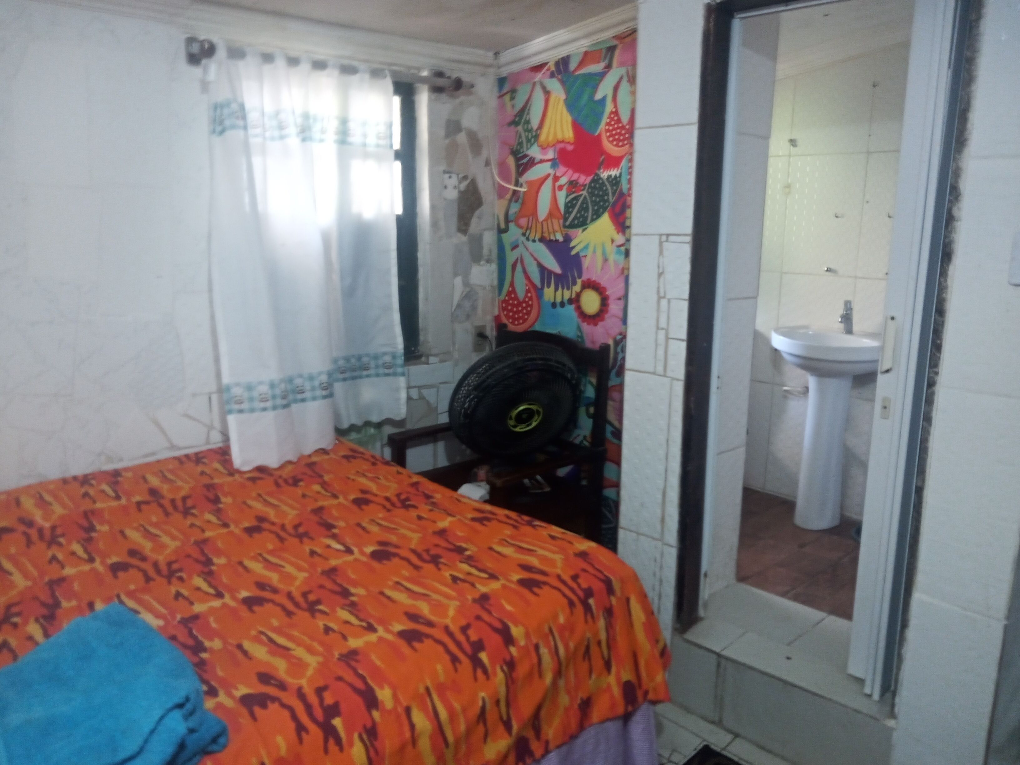 Une chambre à coucher avec un lit, un ventilateur, un rideau et une salle de bain avec un lavabo.