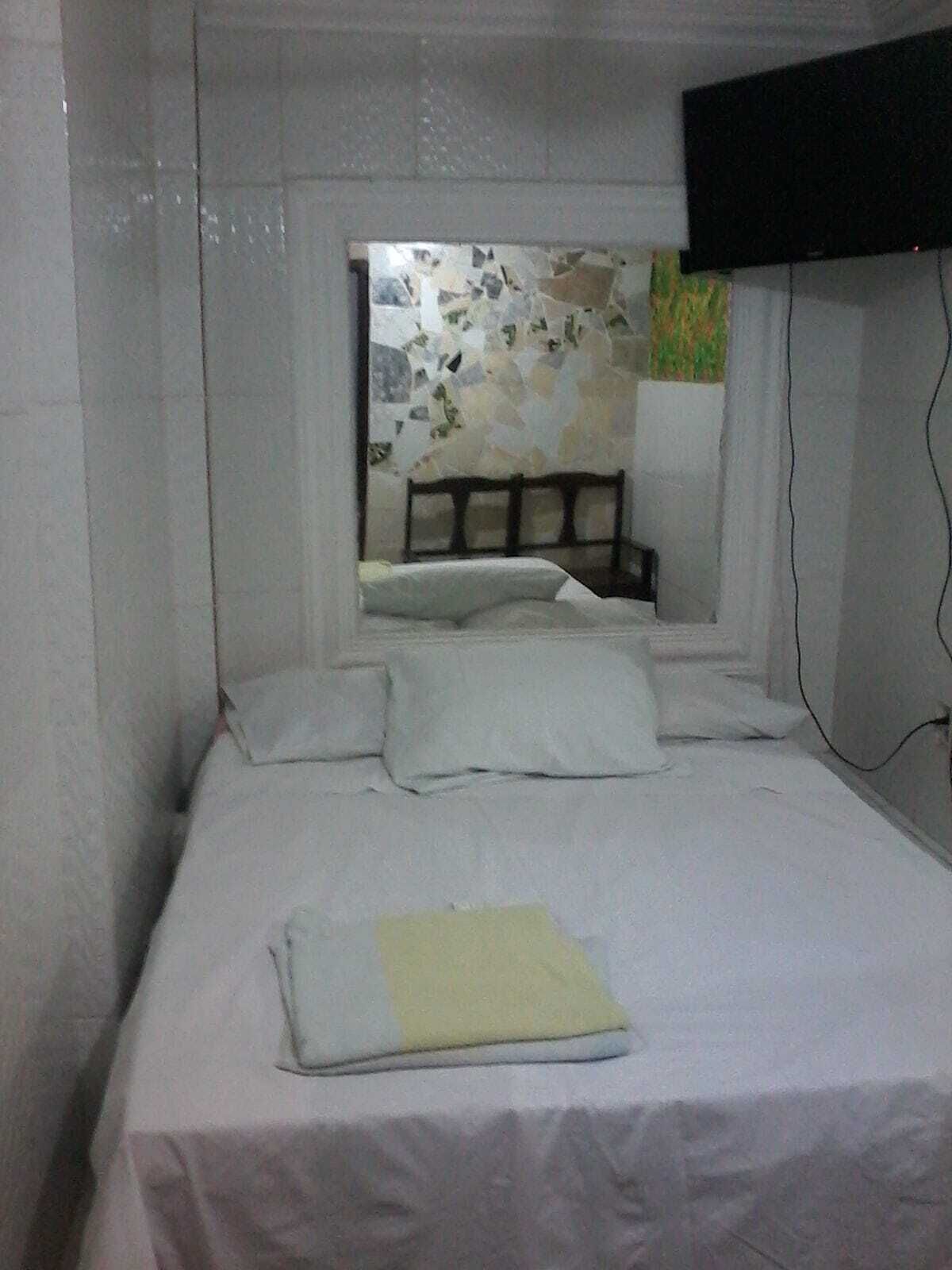 A small, simple room with a bed, a flat-screen TV mounted on the wall, and a window with a view of a building.