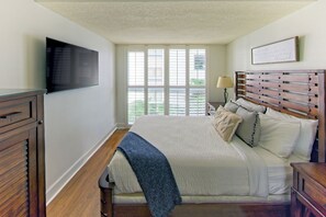 Een slaapkamer met een groot bed, een aan de muur bevestigde televisie, een raam met zonwering en een nachtkastje met een lamp.