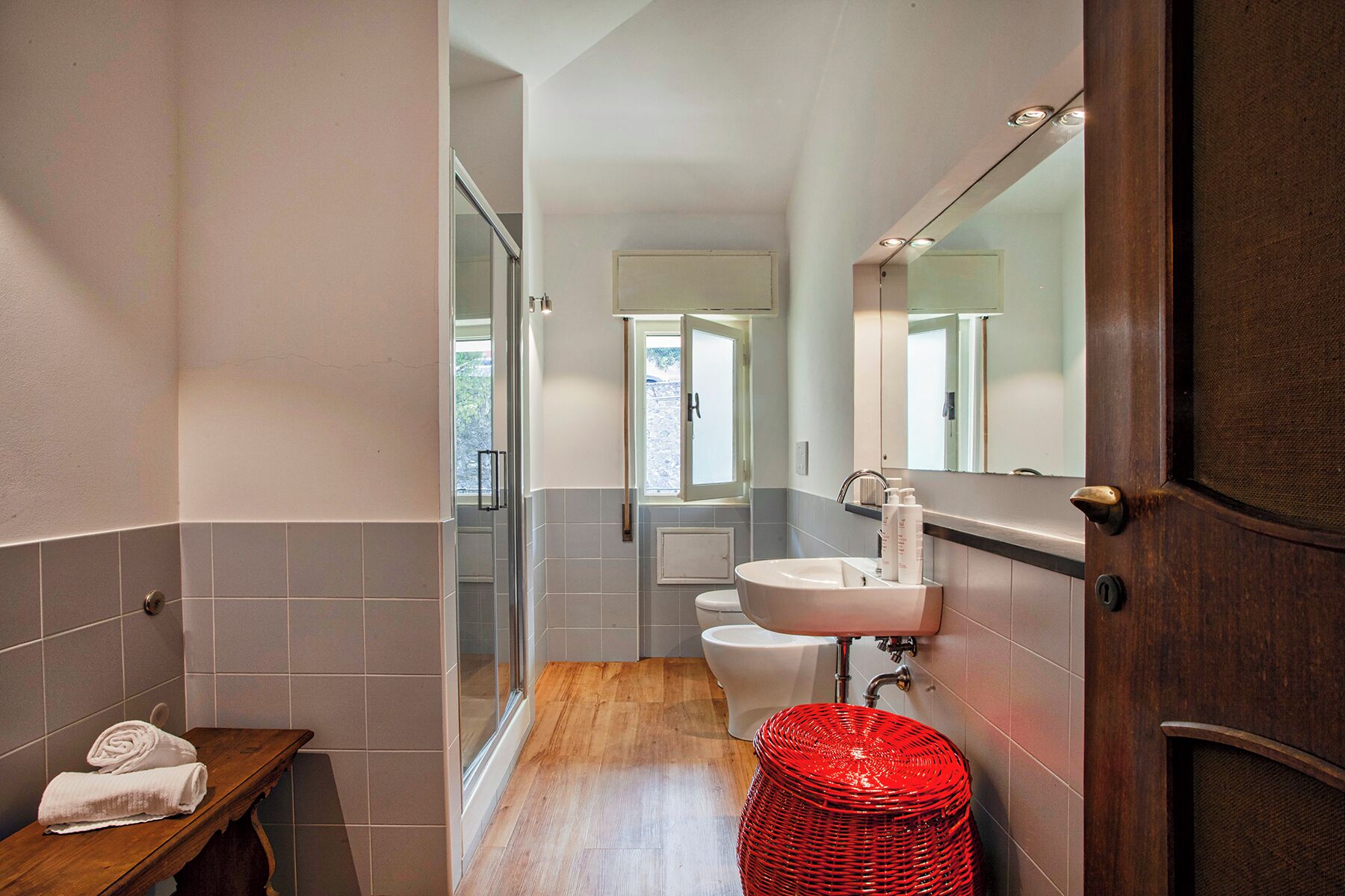 A modern bathroom with a glass shower, a wooden bench, and a red wicker basket.