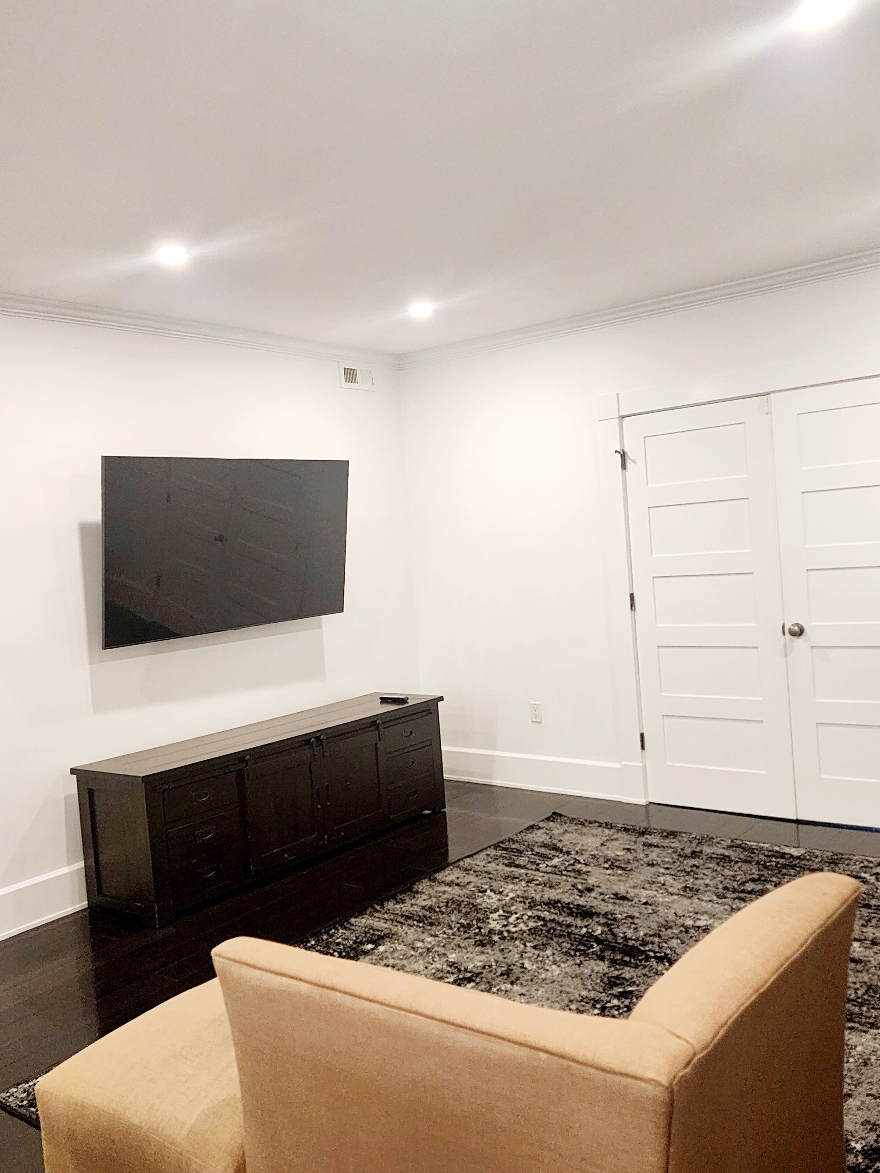 A room with a flat-screen TV mounted on the wall, a dark wooden cabinet, a beige sofa, and a white door.