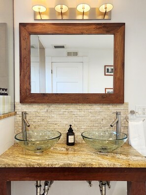 A bathroom with a glass bowl sink, two faucets, a wooden-framed mirror, and a wall-mounted light fixture. - Comfort and Location! (Medford)