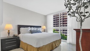 A hotel room with a large bed, two bedside lamps, a nightstand with drawers, a wooden nightstand, and a view of a building through a window with white curtains. - Modern Bay View with Free Parking (Miami)