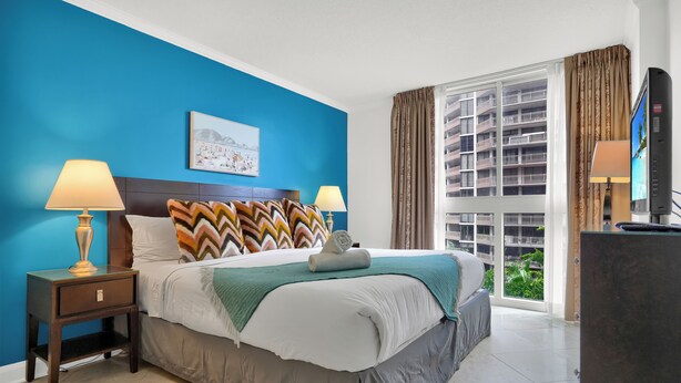 A hotel room with a large bed, two bedside tables with lamps, a window with curtains, and a view of a building. - Relaxing Condo with Free Parking (Miami)