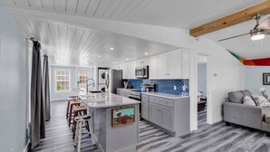 A modern kitchen with white cabinets, a central island with stools, and a dining area with a gray sofa. - Blue Sky Ranch-walk to beach! Sleeps 6 (Murrells Inlet)