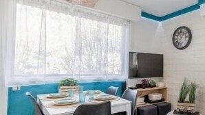Ein Essbereich mit einem weißen Tisch, gedeckt für vier Personen, einem großen Fenster mit durchsichtigen Vorhängen, einer modernen Chandelierleuchtre und einem an der Wand montierten Flachbildfernseher.
