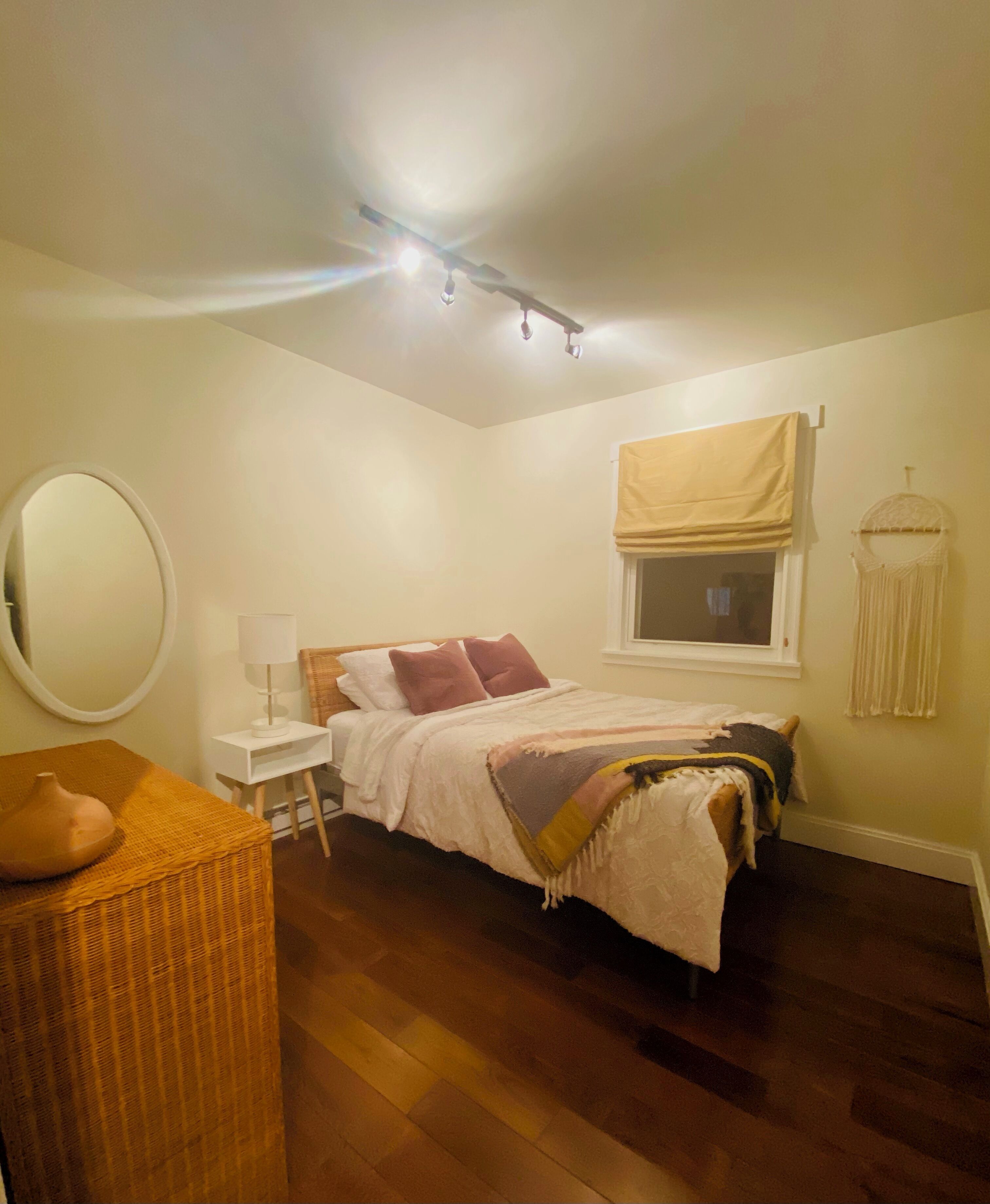 Une chambre à coucher comprenant un lit, une table de chevet, une lampe, une fenêtre avec un store, un luminaire suspendu et un miroir.