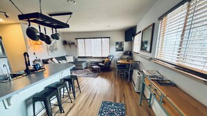 A modern kitchen with a bar area, wooden flooring, and a large window with blinds.
