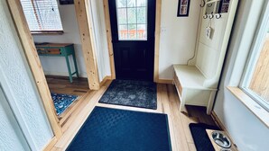 A hallway with a wooden ceiling, a door leading to a room, a bench, a coat rack, and a window with blinds.