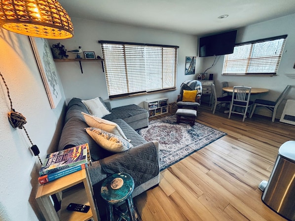 Een woonkamer met een bank, een bijzettafeltje met boeken, een stoel, een televisie en een raam met jaloezieën.