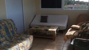 A room with a sofa, a small table, a wicker chair, a window with a view, and a hanging light fixture. - Home. (Zonza)