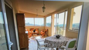 A room with a dining table, chairs, a wooden cabinet, and a view through large windows. - Home. (Zonza)