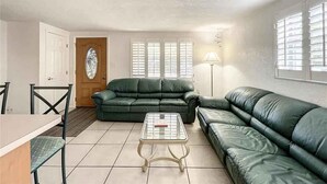 A living room with a green sofa, a coffee table, and a lamp. There is a door and windows with blinds. - Kick back and Relax at Leisure Beach (Hudson)