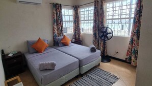 A bedroom with two beds, a standing fan, and a window with decorative curtains.