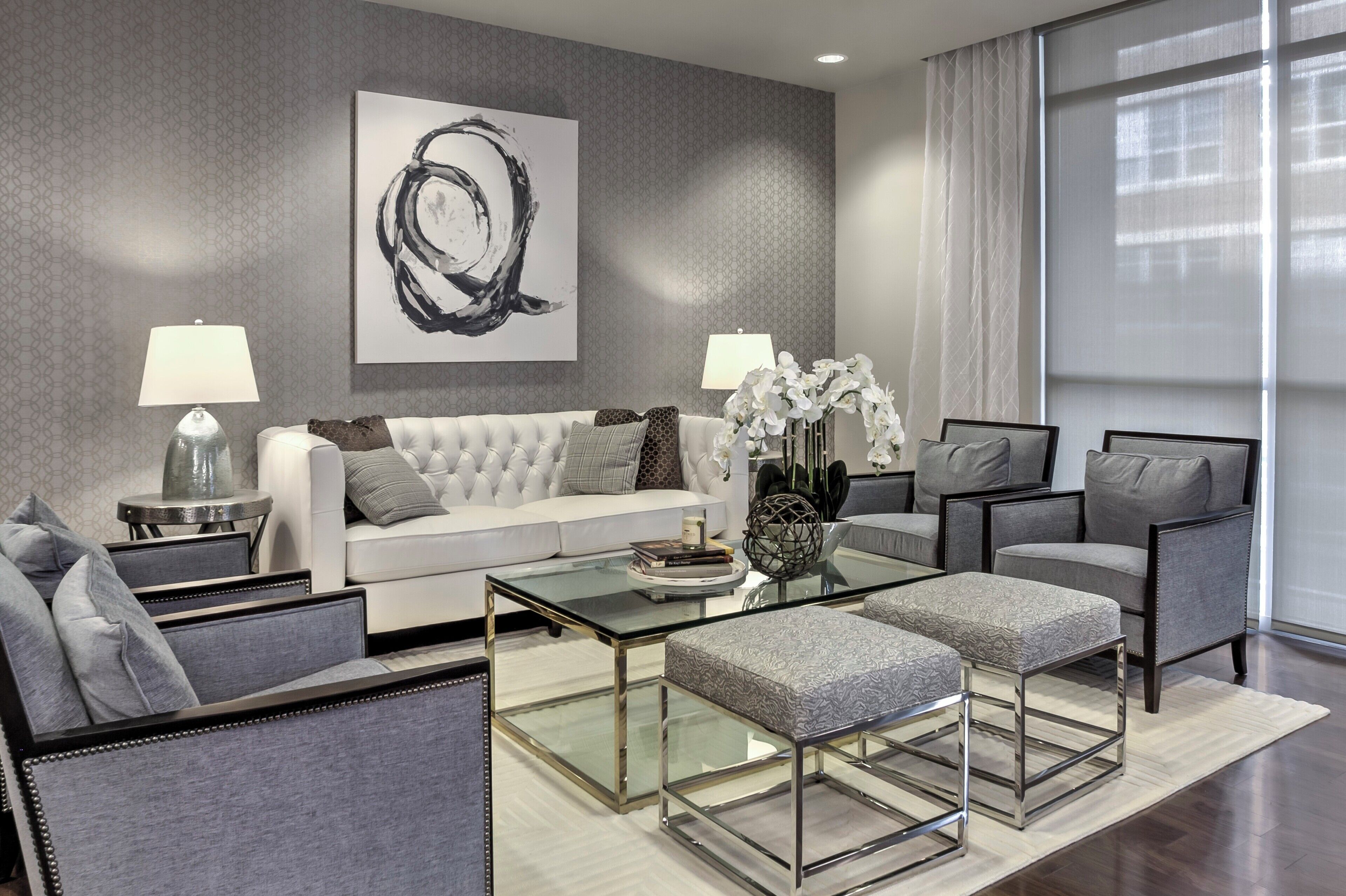 A modern living room with a white sofa, grey armchairs, a glass coffee table, and a large abstract painting on the wall.