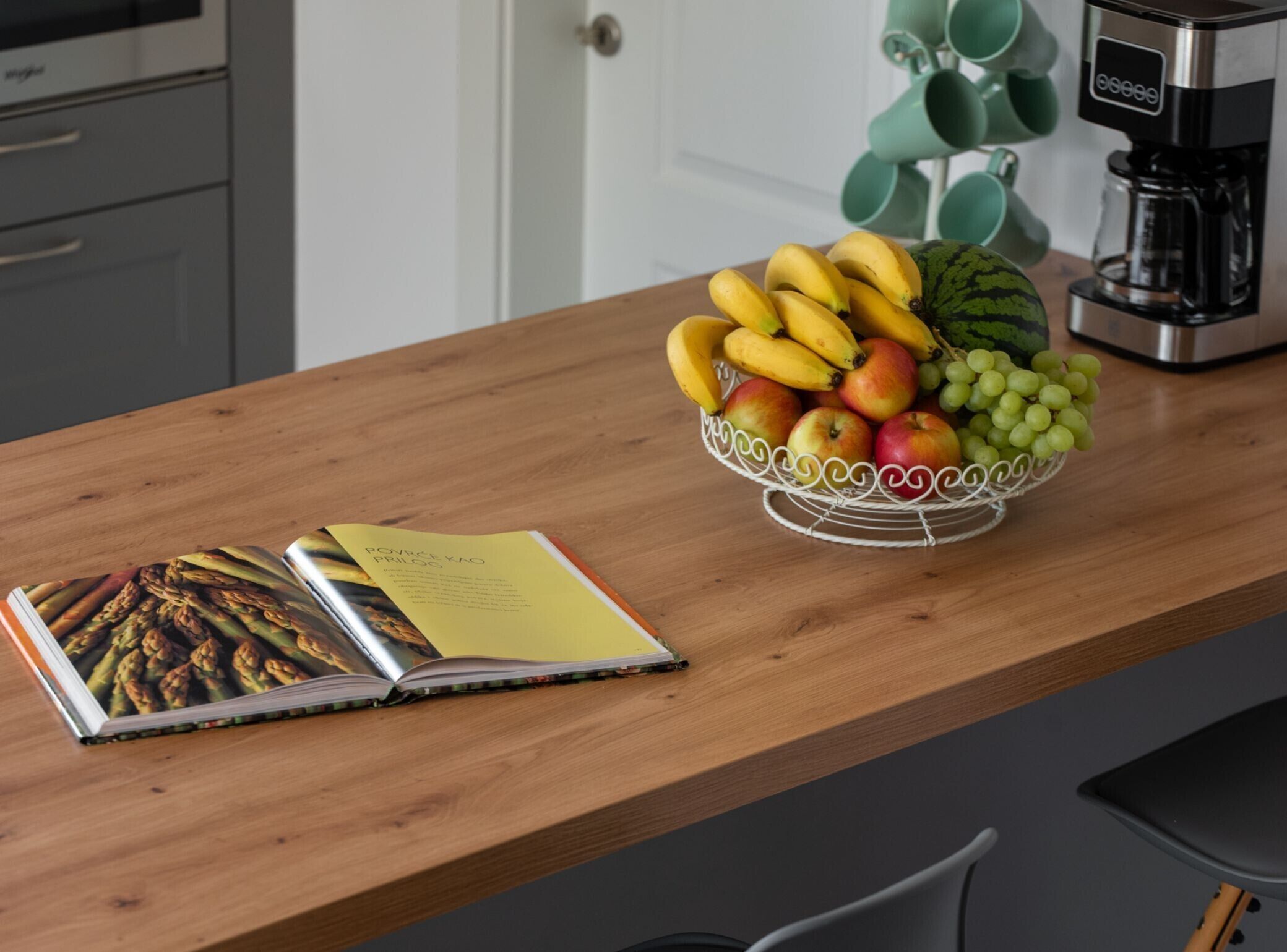 Un comptoir de cuisine avec un panier de fruits contenant des bananes, des pommes et des raisins, une machine à café et un livre ouvert dont une page illustre des asperges.