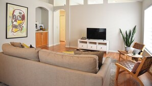 Una sala de estar con un sofá, un televisor sobre un mueble blanco, una planta y un ventilador de techo.
