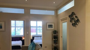 Ein geräumiges Wohnzimmer mit einer großen Couch, einem Couchtisch und einem Bücherregal. Im Hintergrund sind zwei Betten, eine Tür und ein Fenster mit Jalousien zu sehen.