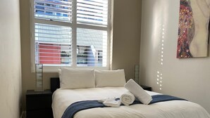 Ein Hotelzimmer mit Bett, Fenster mit Jalousien, Nachttisch und einem Gemälde an der Wand.