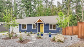 A single-story house with a blue exterior and a yellow front door, surrounded by a gravel driveway and small plants. - Freeland Retreat - Deck, Fire Pit & Beach Access! (Freeland)
