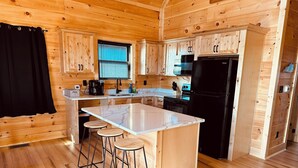 A wooden cabin interior with a kitchen area featuring a large island with stools, wooden cabinets, and a black refrigerator.