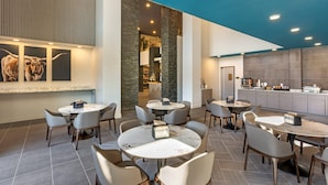 A modern dining area with round tables and chairs, a kitchen in the background, and a wall with a bull painting.