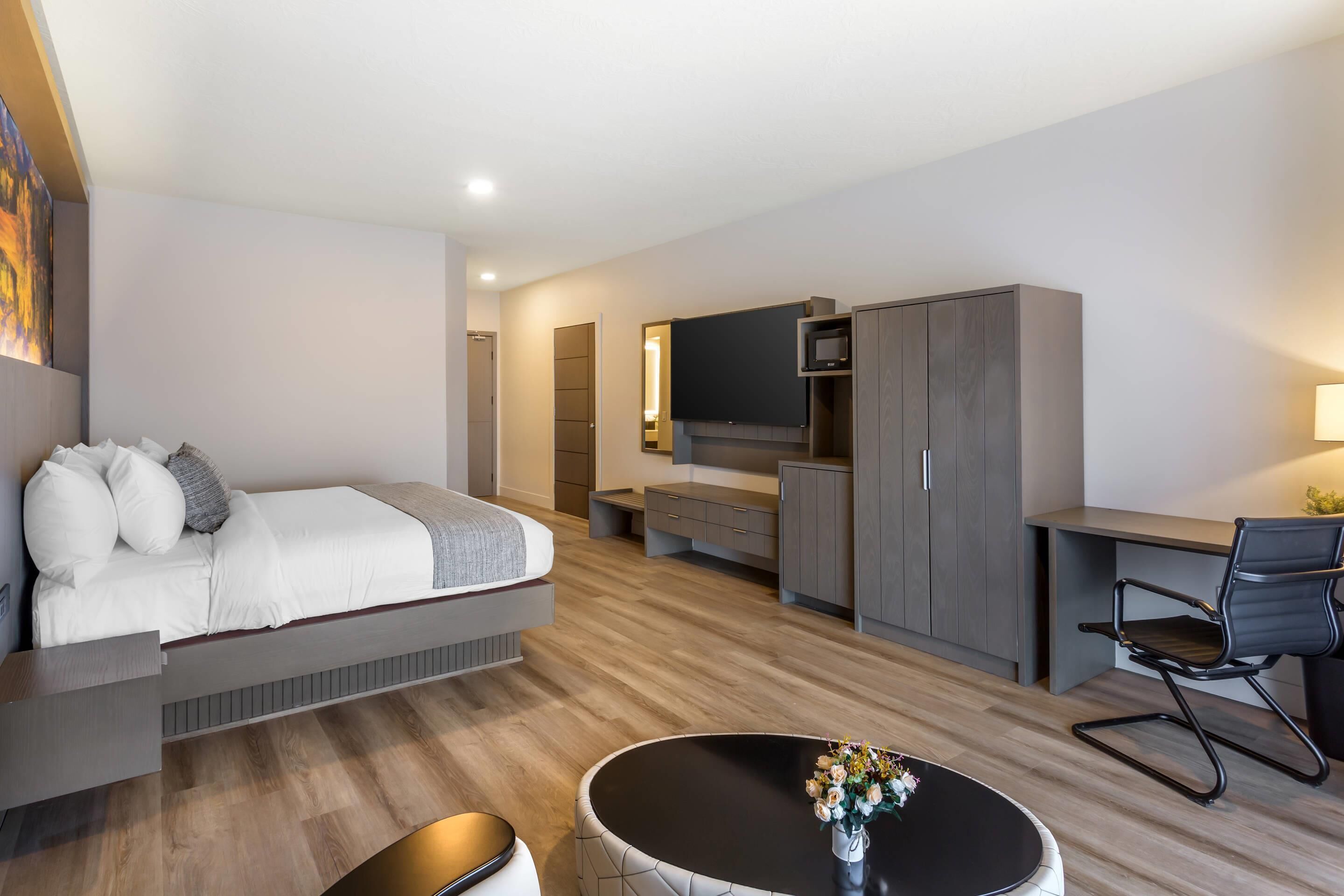Ein modernes Hotelzimmer mit Bett, Schreibtisch, Stuhl, Fernseher, Kleiderschrank und einem kleinen runden Tisch mit Blumen.