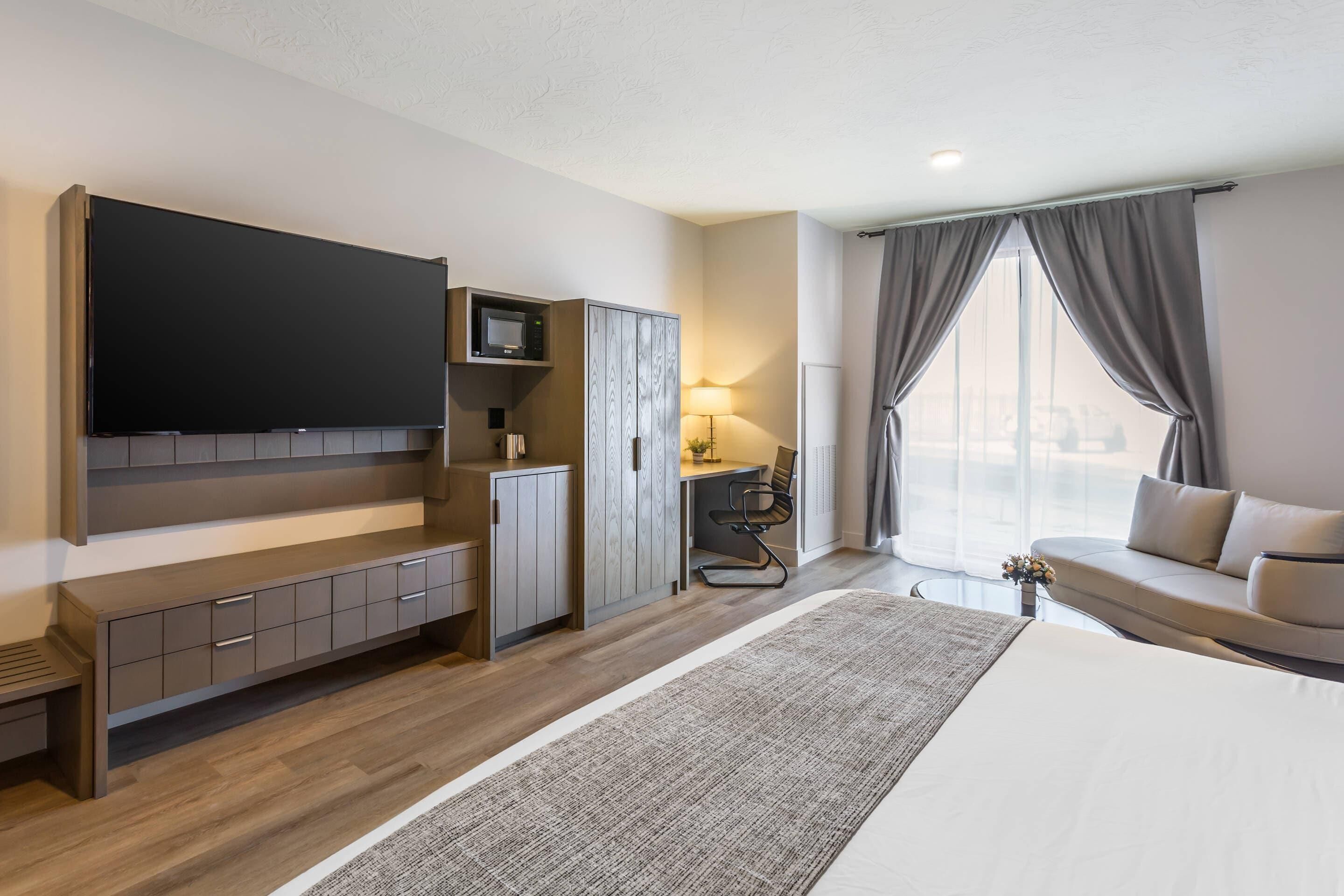 A modern hotel room with a large flat-screen TV, a desk with a lamp, a sofa, and a large window with curtains.