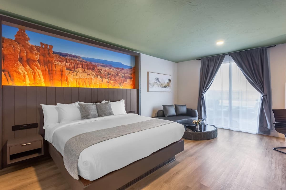 A modern hotel room with a large bed, a wall-mounted TV displaying a scenic landscape, a seating area with a round table, and a large window with curtains.