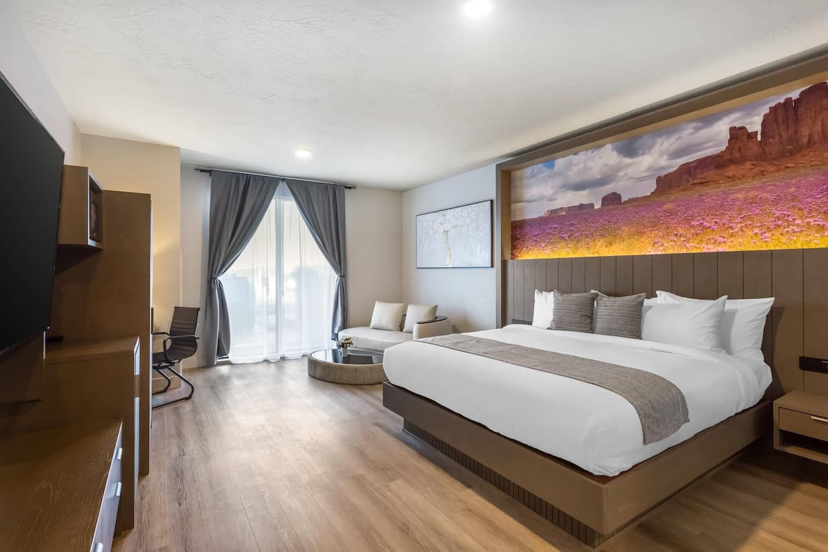 A modern hotel room with a large bed, a flat-screen TV, a seating area with a sofa and chair, and a painting of a desert landscape on the wall.
