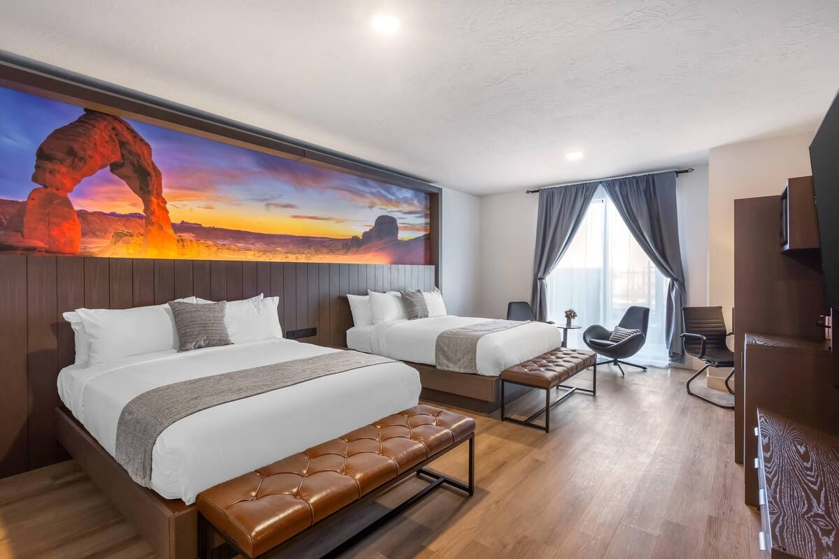 A hotel room with two beds, a bench, a desk, a chair, and a large painting of a rock formation.
