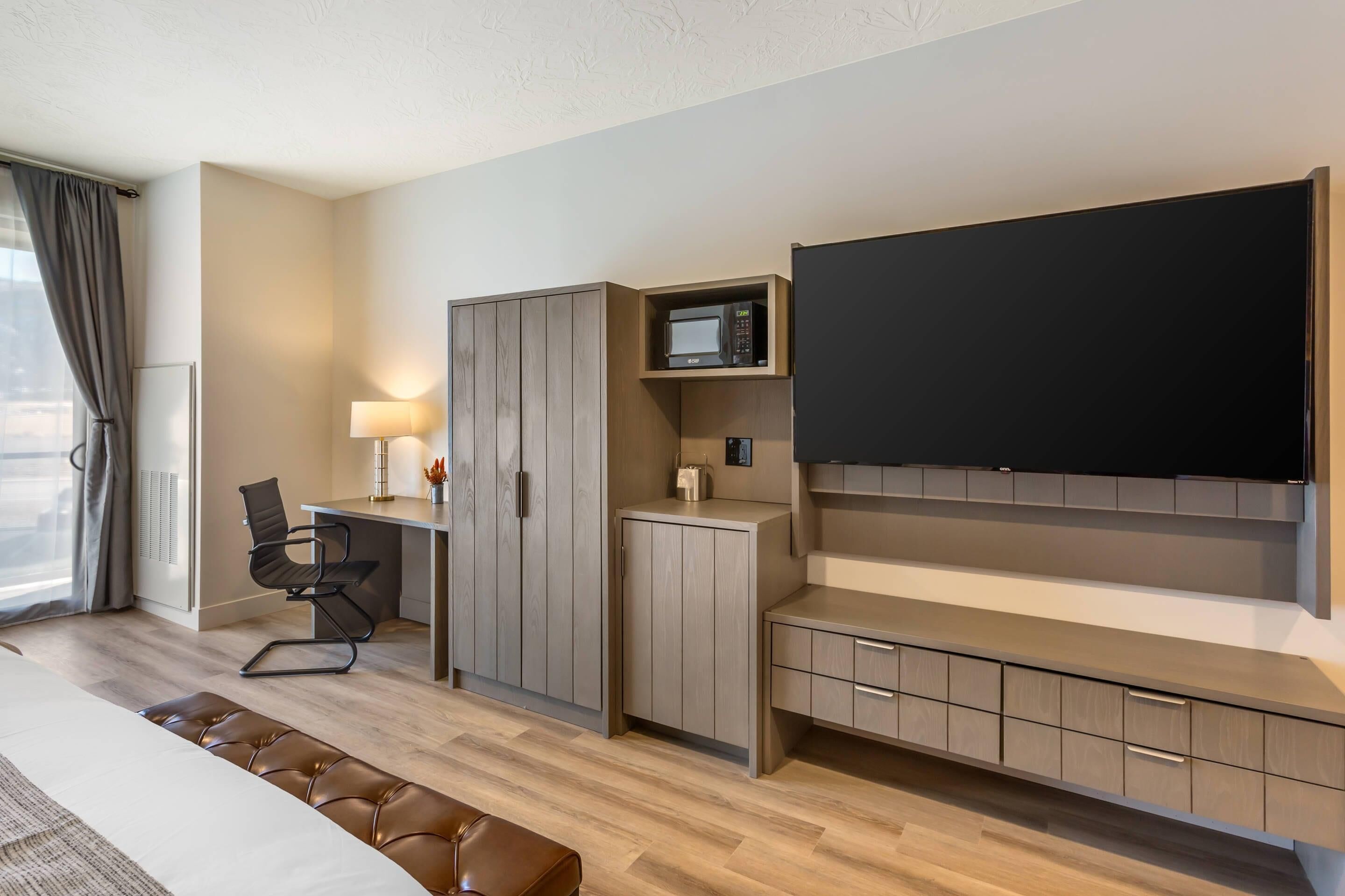 A modern hotel room with a flat-screen TV, a desk with a lamp, a chair, a wardrobe, and a bench.