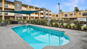 Une piscine dotée d’un parasol, entourée d’une clôture, et située à côté d’un immeuble à plusieurs étages avec des balcons.