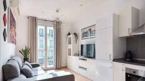 Una sala moderna con un sofá gris, un televisor y una zona de cocina con armarios blancos.