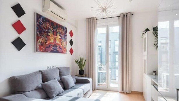 Una sala de estar moderna con un sofá gris, un cuadro abstracto colorido y un amplio ventanal con cortinas.