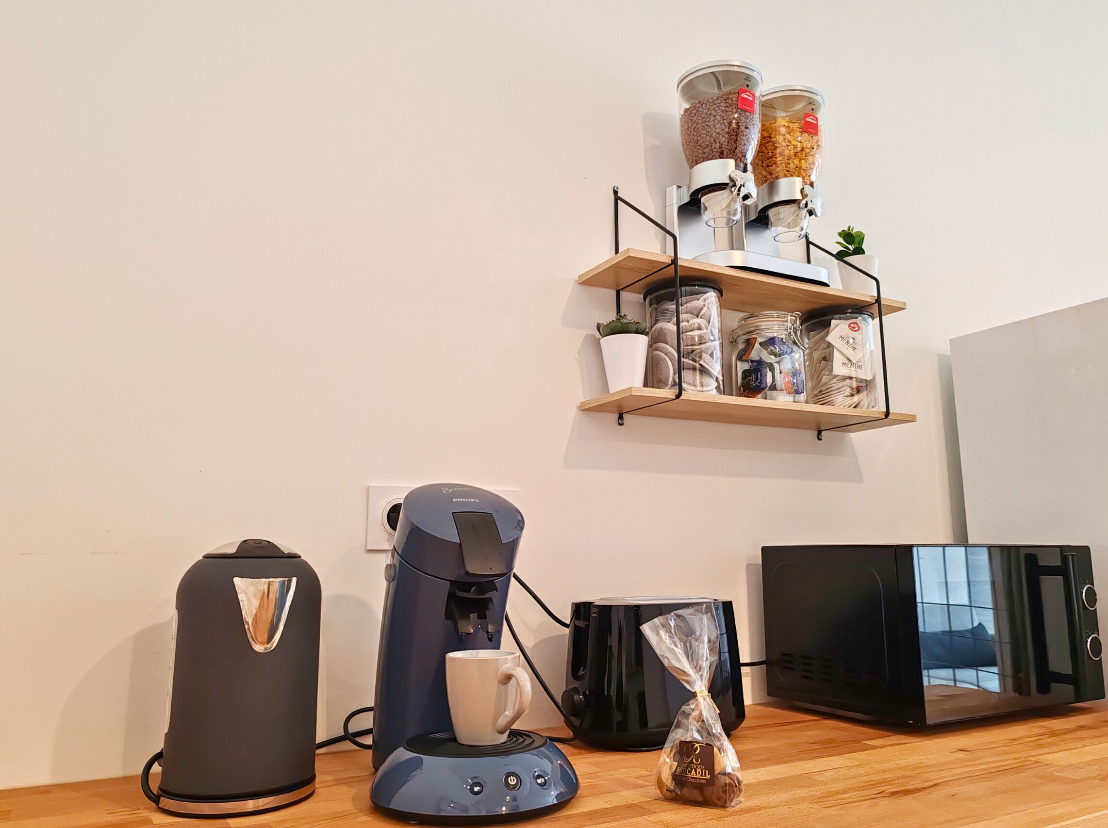 A kitchen with a coffee maker, a microwave, a toaster, and a shelf with jars and a potted plant.
