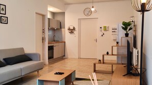 A modern living room with a sofa, coffee table, and TV stand. There is a kitchen in the background with cabinets and a refrigerator. - Chill and Work  (Villeneuve-sur-Lot)